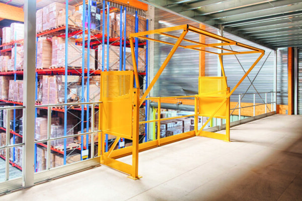 Sas de sécurité jaune sur une plateforme élevée dans un entrepôt industriel, entouré d’étagères bleues remplies de marchandises