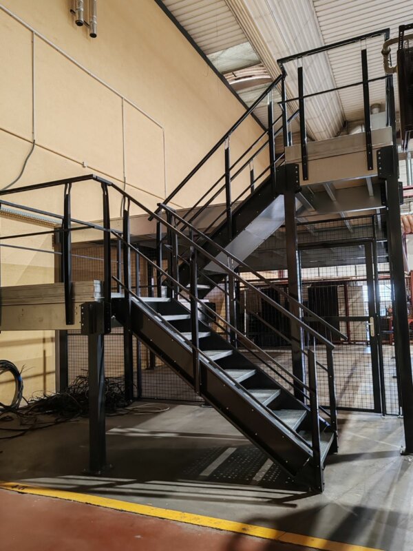 Il s’agit d’une photo d’un escalier métallique noir dans un entrepôt. L’escalier est en métal noir et a un palier à mi-chemin. L’escalier est contre un mur beige et a une rambarde noire. L’escalier mène à un deuxième étage avec un sol en grille métallique. L’arrière-plan est un entrepôt avec un sol en béton et des étagères métalliques.