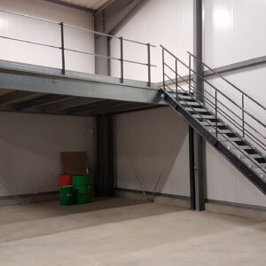 Photo d’une mezzanine métallique avec des escaliers dans un entrepôt. La mezzanine est en métal gris et a une rambarde autour du niveau supérieur. Les escaliers sont situés sur le côté droit de la mezzanine et mènent au niveau supérieur. Il y a quelques boîtes et barils sur le niveau inférieur de la mezzanine. L’arrière-plan est un mur d’entrepôt blanc avec une grande porte sur le côté gauche.