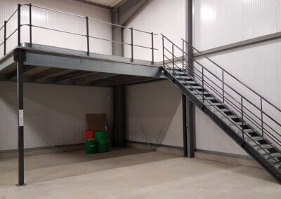 Photo d’une mezzanine métallique avec des escaliers dans un entrepôt. La mezzanine est en métal gris et a une rambarde autour du niveau supérieur. Les escaliers sont situés sur le côté droit de la mezzanine et mènent au niveau supérieur. Il y a quelques boîtes et barils sur le niveau inférieur de la mezzanine. L’arrière-plan est un mur d’entrepôt blanc avec une grande porte sur le côté gauche.